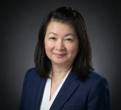 Professional studio headshot of a smiling woman with shoulder-length dark hair, wearing a navy blue blazer over a white blouse against a dark gray gradient background.
